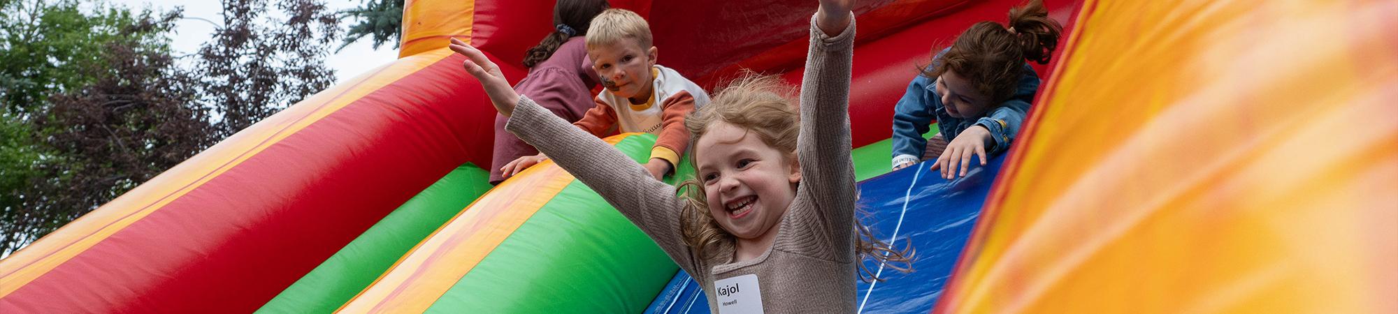 A young child on a bouncy slide with her arms up and smiling as 